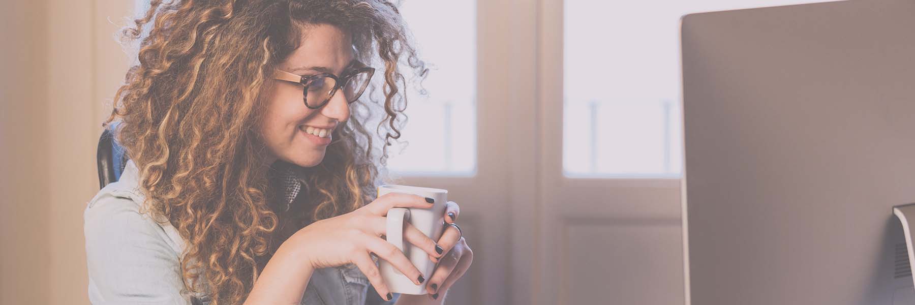 A woman smiling on her computer holding a white mug